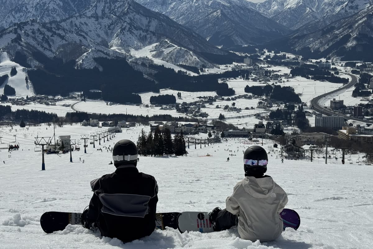 兩位單板玩家並肩坐在雪地上，背對鏡頭眺望遠方城鎮與雪山