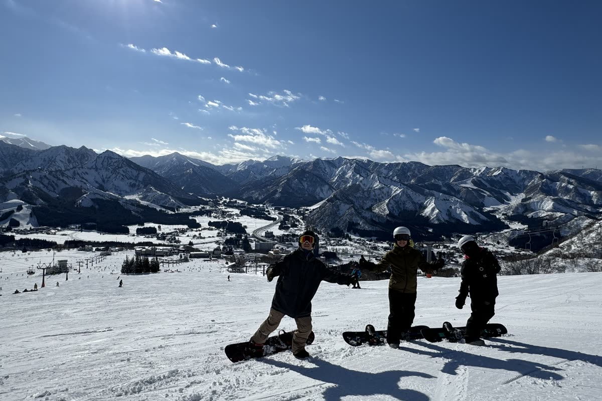 三位單板玩家站在岩原的雪道頂端張開雙手，背景是湯澤市街與群山