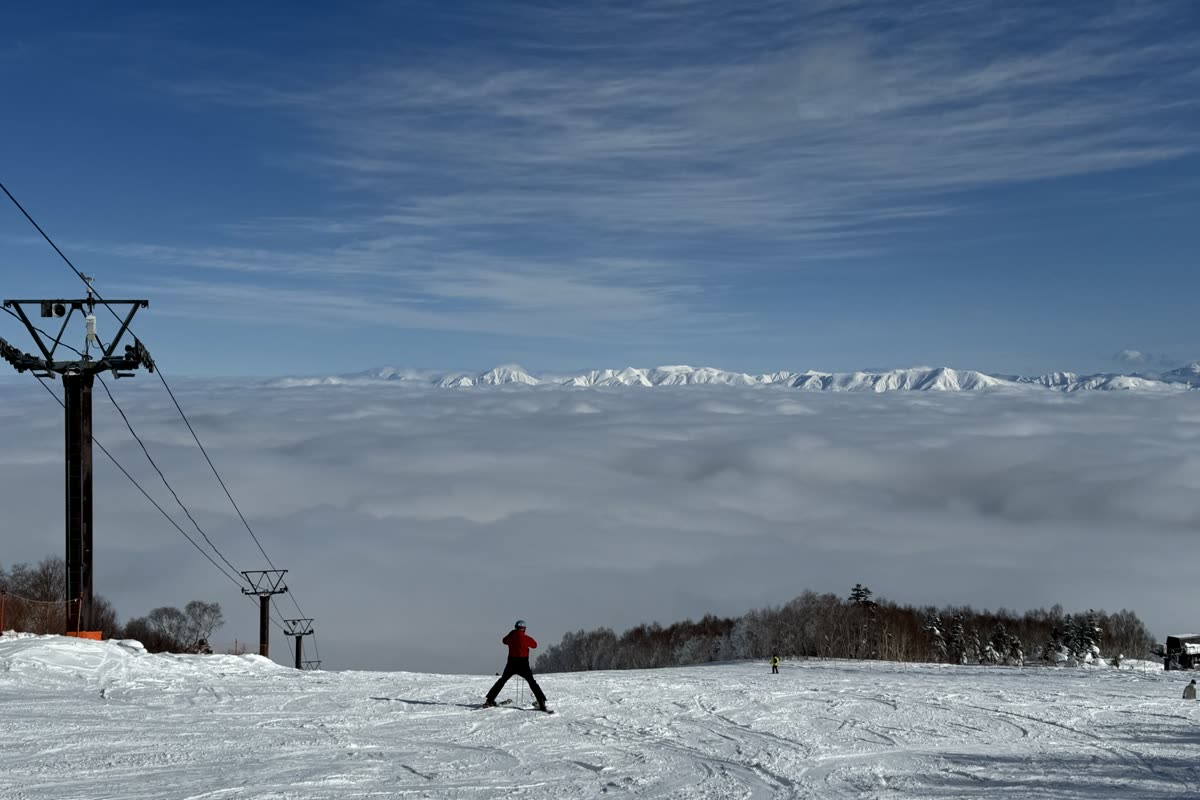 三位滑雪與單板玩家在雪道中段轉彎下滑，背景是田代湖與遠山