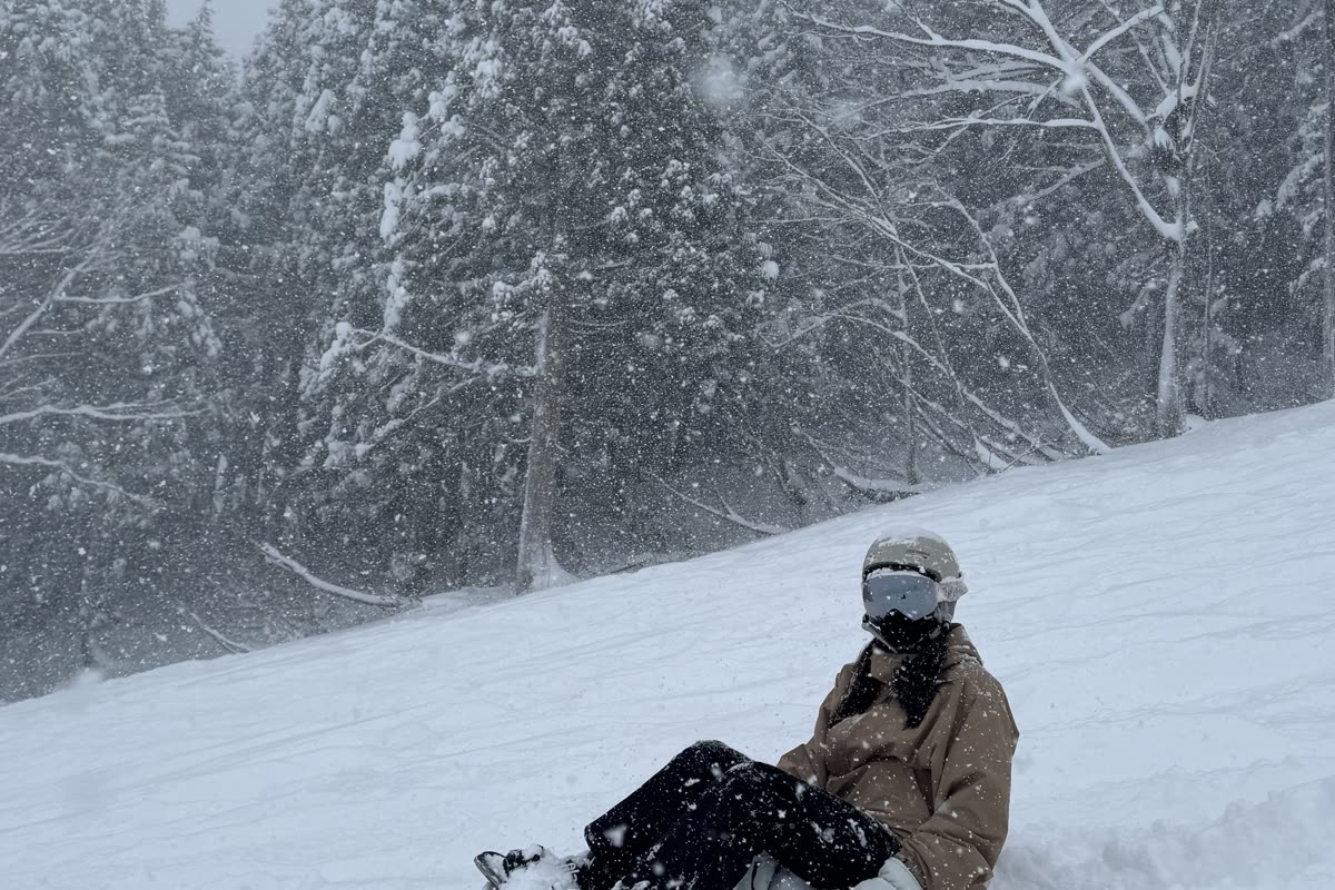 大雪中一位戴著雪鏡與面罩的滑雪者坐在雪地上，背景是針葉林
