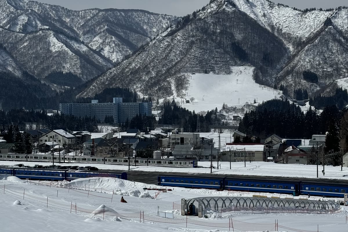 從雪場遠眺越後中里站方向，可見停靠的藍色電聯車、隧道與雪山